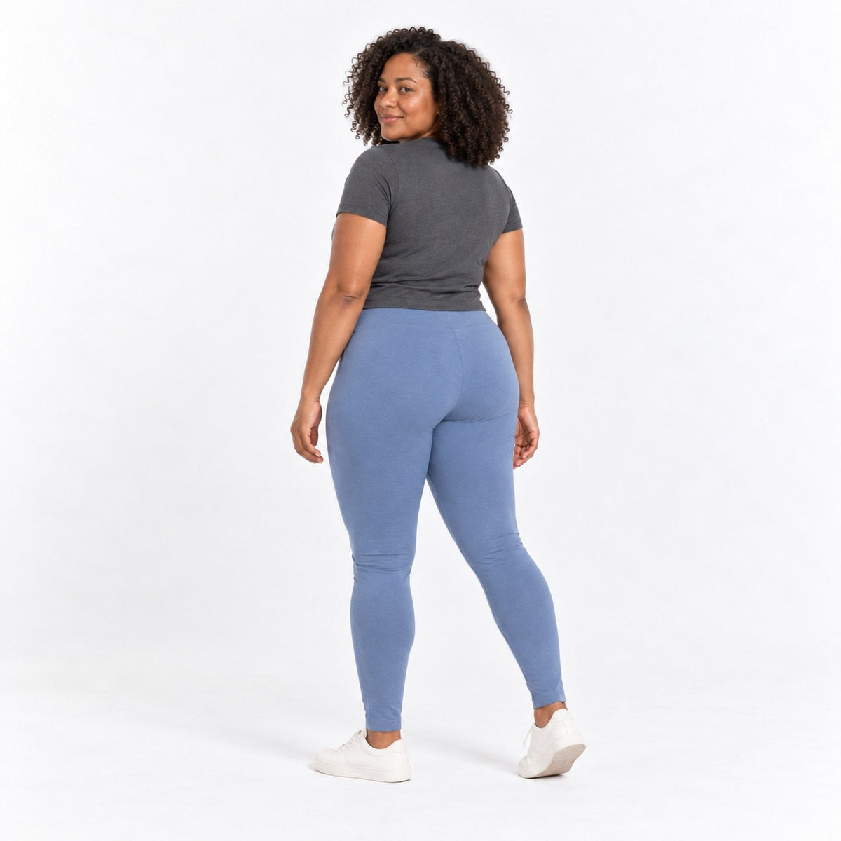 A woman is shown from the back wearing a dark gray short-sleeve t-shirt, dusty blue organic cotton high waist leggings , and white sneakers against a plain white studio background.