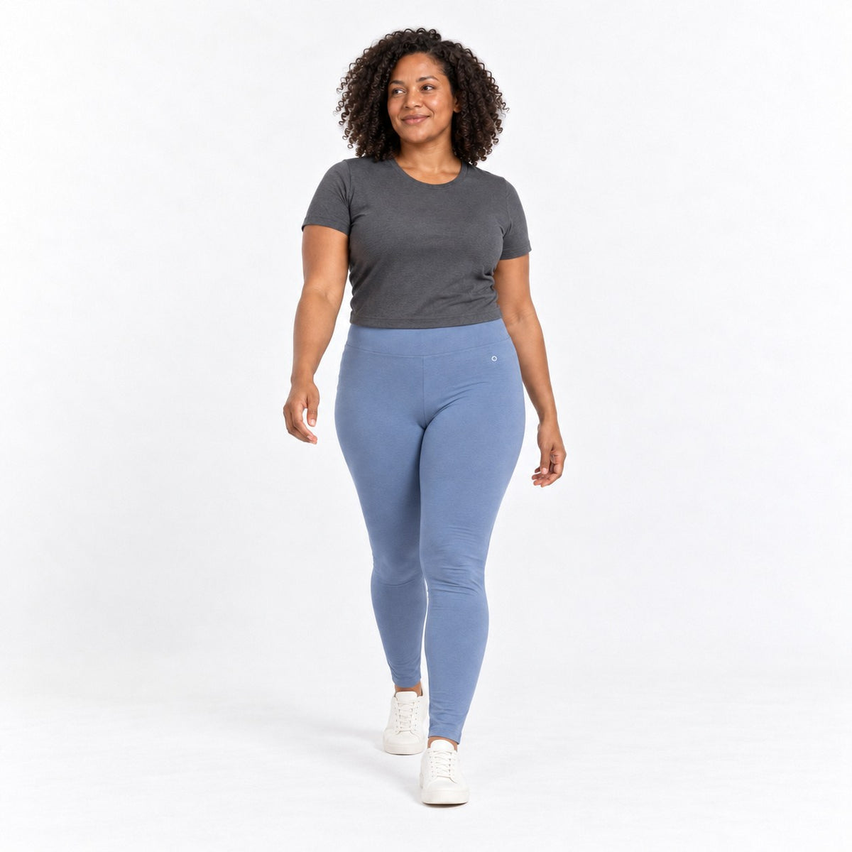A woman modeling a dark gray short-sleeve t-shirt and light blue organic cotton high waist leggings with white sneakers, walking against a plain white studio background.