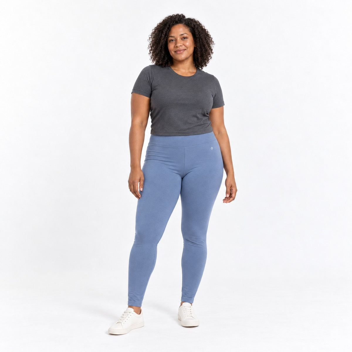 A woman modeling a dark gray short-sleeve t-shirt and light blue h organic cotton high waist leggings with white sneakers, standing against a solid white studio background.