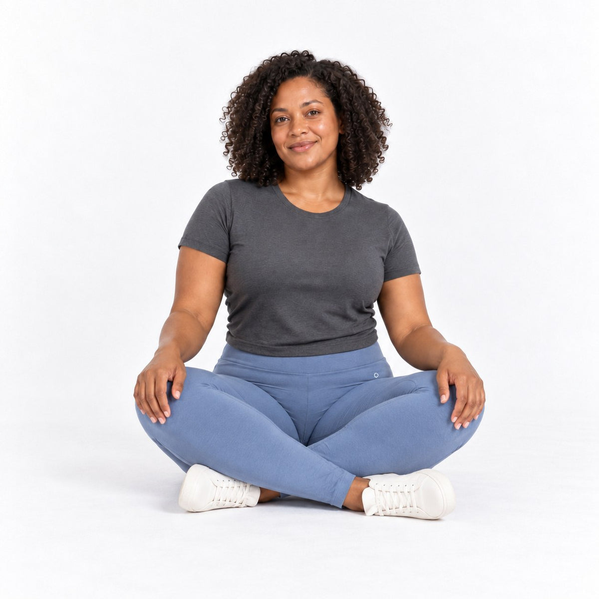 A woman with curly hair sits cross-legged on a white studio background, wearing a dark gray short-sleeve t-shirt, slate blue organic cotton high waist leggings , and white sneakers.