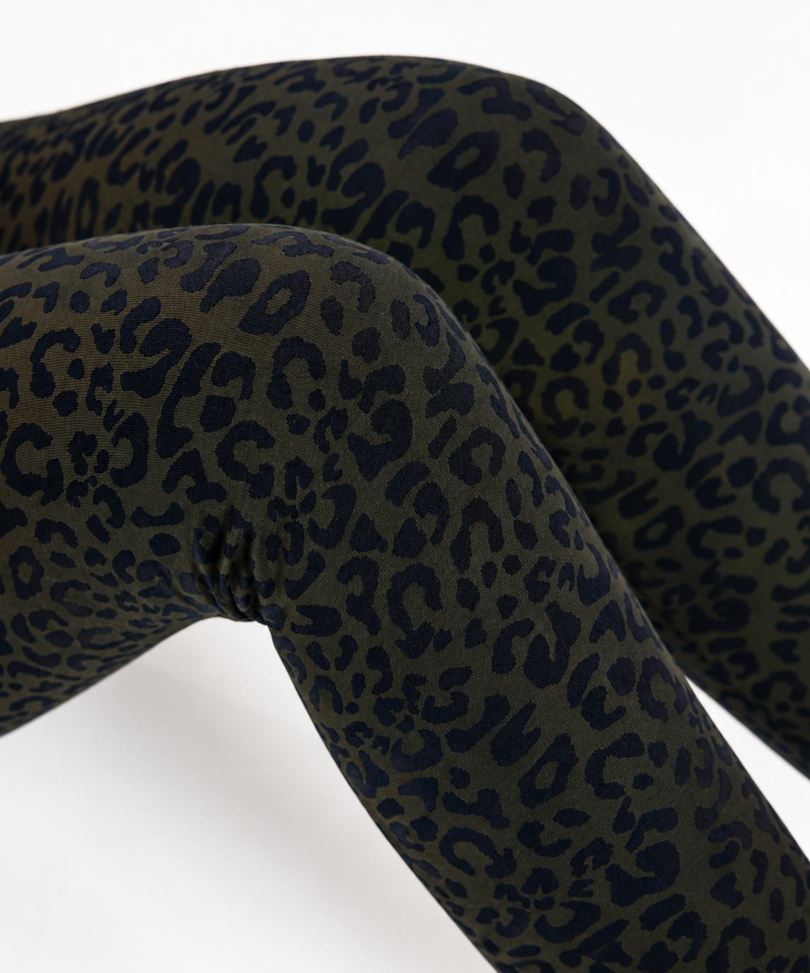 Close-up of leopard-print leggings worn on legs, featuring an olive green base with dark navy spots, set against a plain white background.