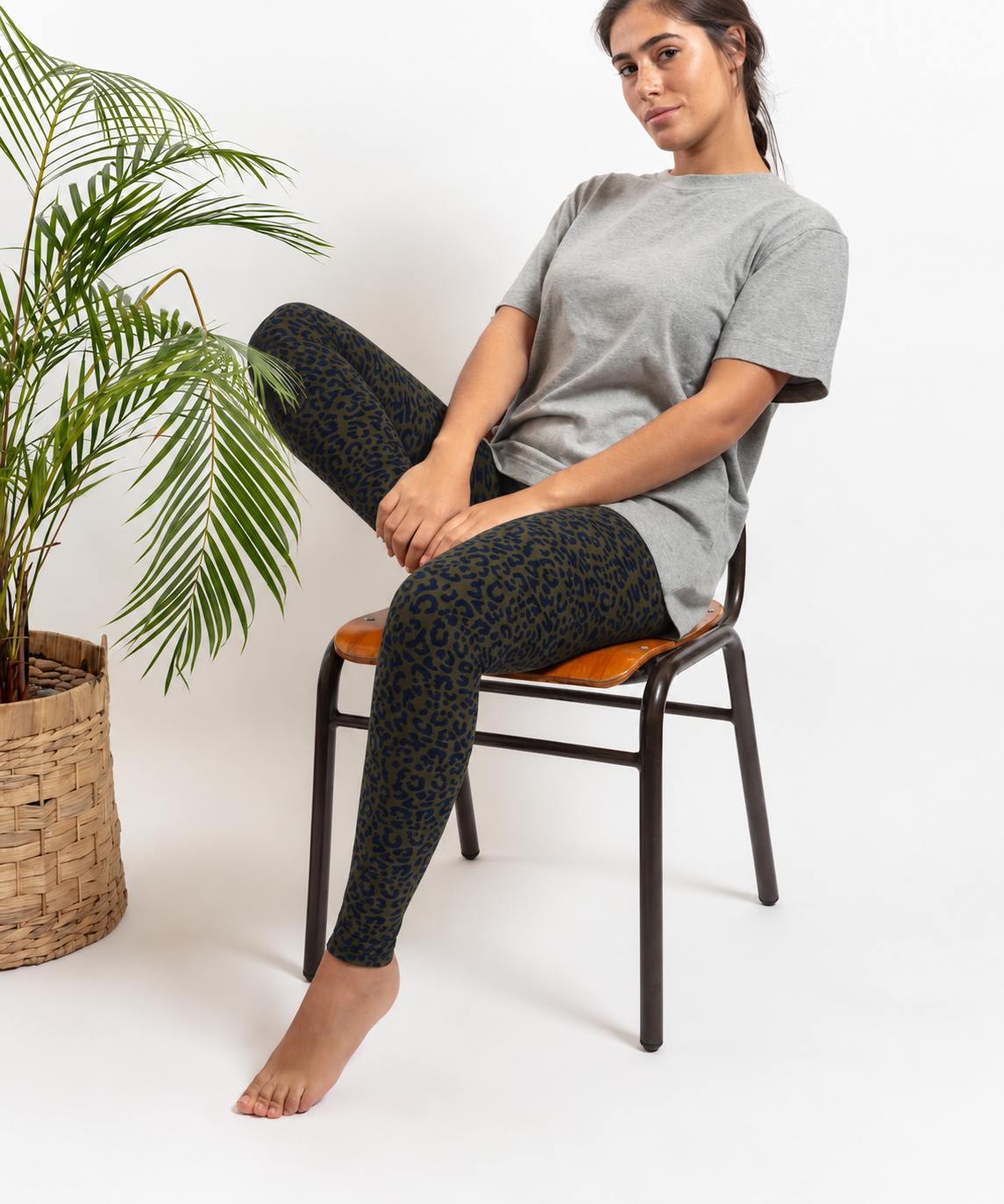 A woman sits on a black chair with a brown cushion, wearing green leopard print leggings and a light gray t-shirt. A potted palm is on the left against a white studio background.