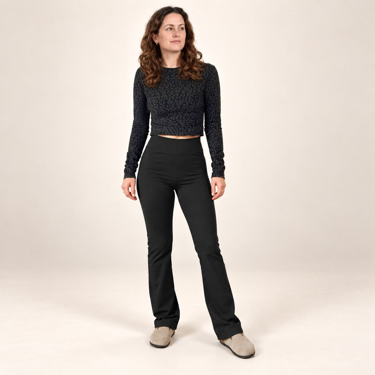 A woman modeling a black textured long-sleeve crop top and black high-waisted flared pants, paired with light gray slip-on shoes, against a plain beige studio background.