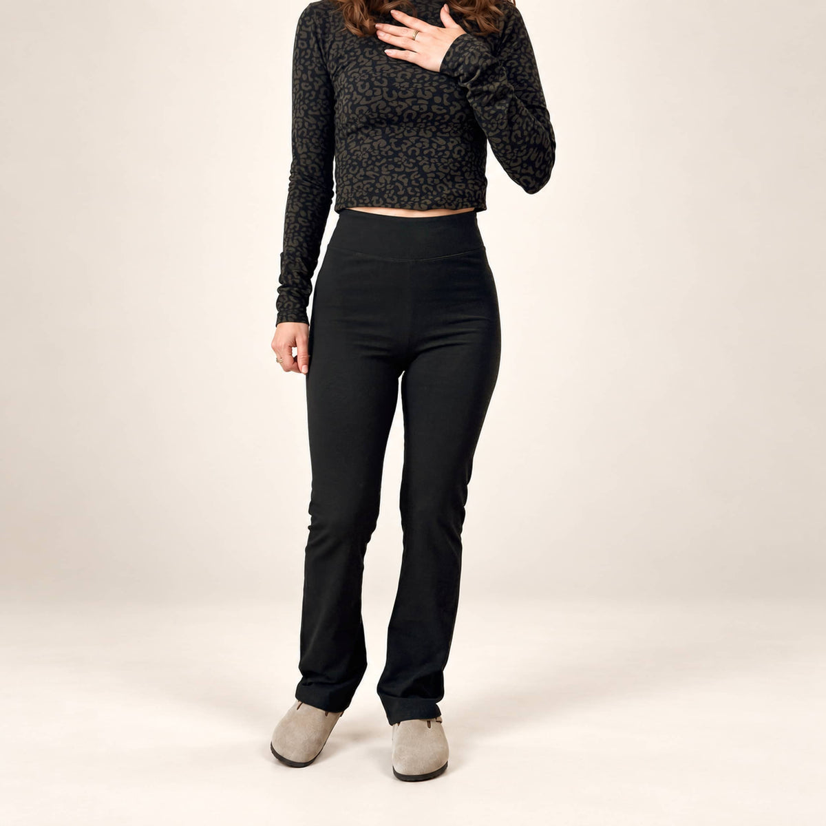 A model wearing black high-waisted k high-waisted organic leggings flared leggings, a black patterned long-sleeve crop top, and gray slip-on shoes, standing against a plain light beige studio background.