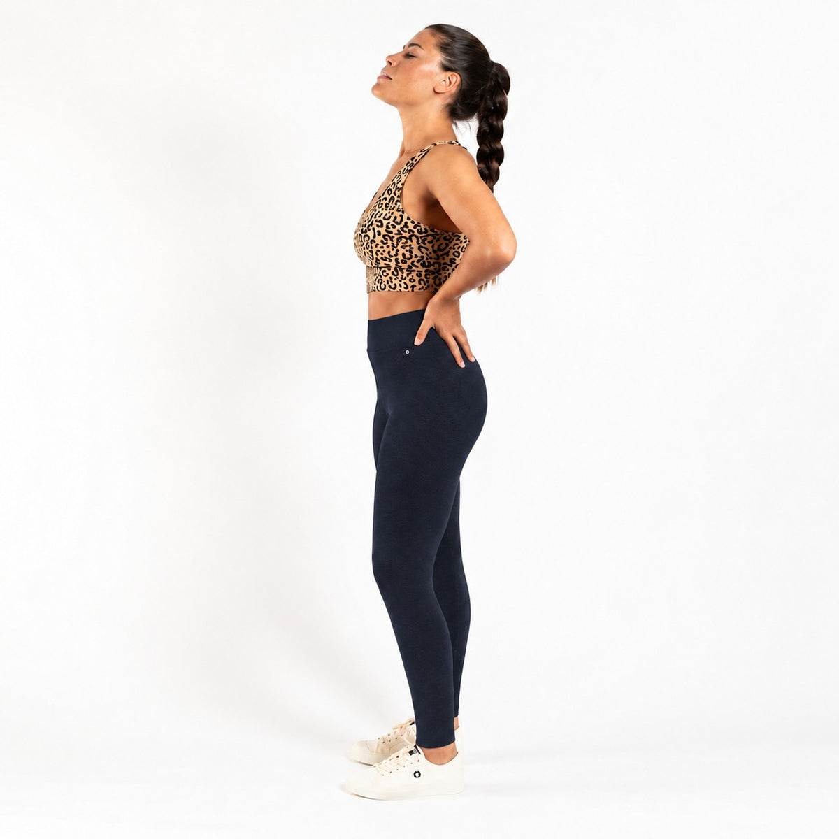 A woman in a side profile pose wearing a leopard print sports bra and navy blue leggings, standing against a plain white studio background.