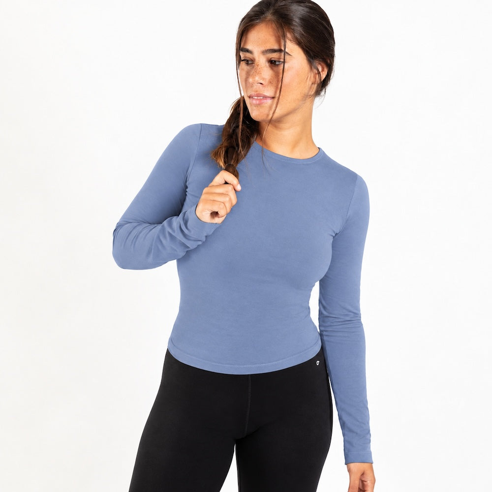 A woman modeling a fitted, organic cotton long sleeve shirt in dusty blue, paired with black leggings, against a plain white studio background.