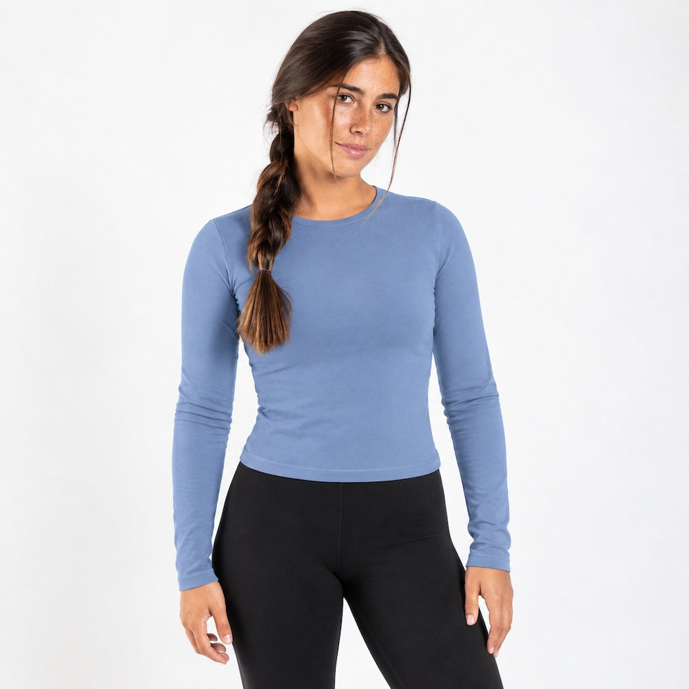 A woman modeling a fitted, organic cotton long sleeve shirt in dusty blue, paired with black leggings, against a plain white studio background.
