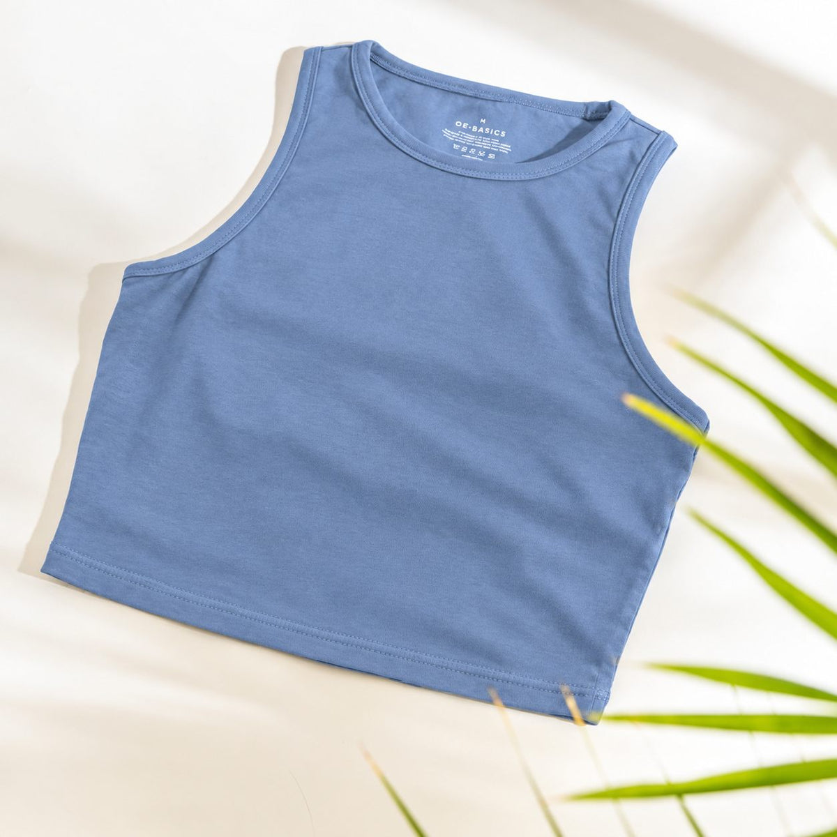 A sleeveless cropped Organic Cotton Tank Top with built in bra in dusty medium blue, laid flat on a light cream background with soft shadows and green plant leaves at the edge.