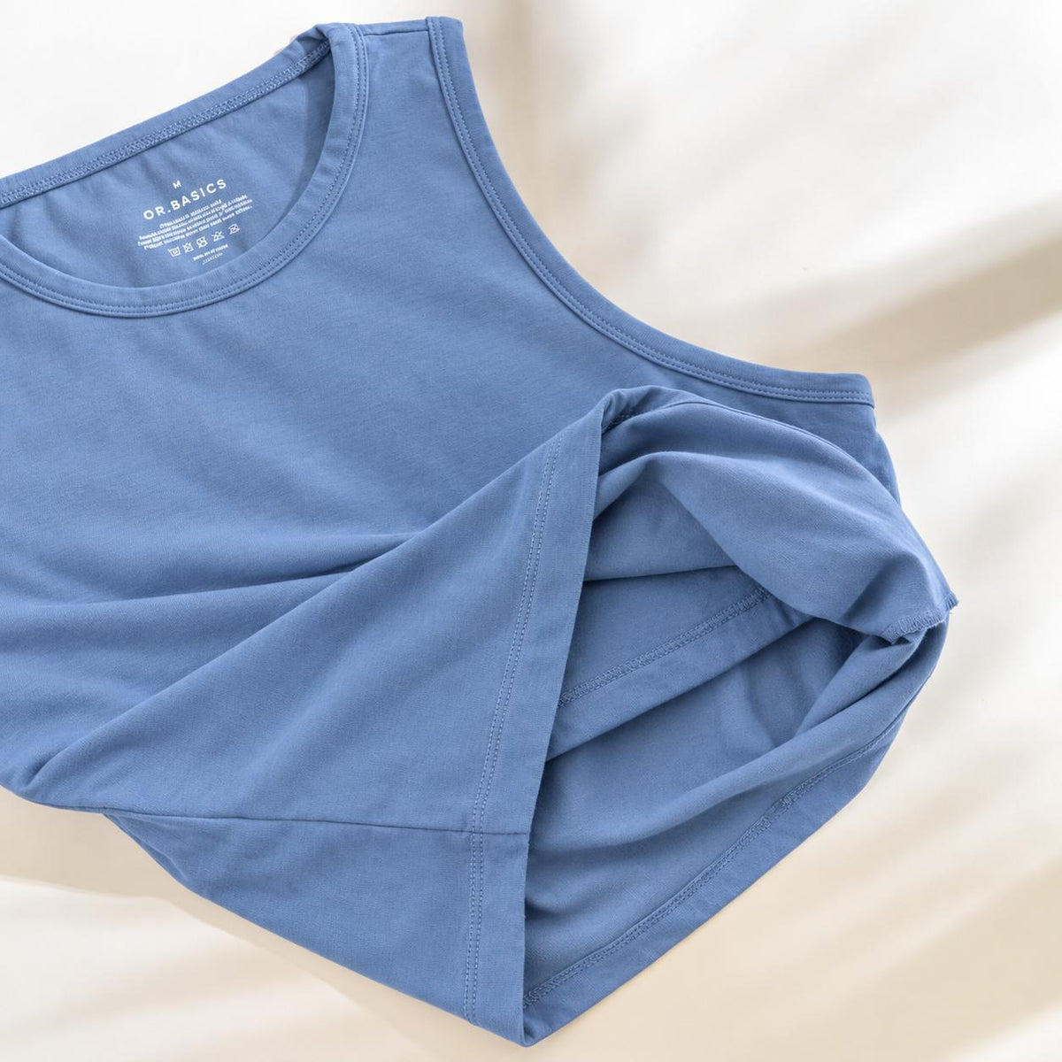 A folded light blue Organic Cotton Tank Top with built in bra with a crew neck, laid flat on a soft beige fabric background. The brand name 'ORIGAMICS' is visible on the inner neck label.