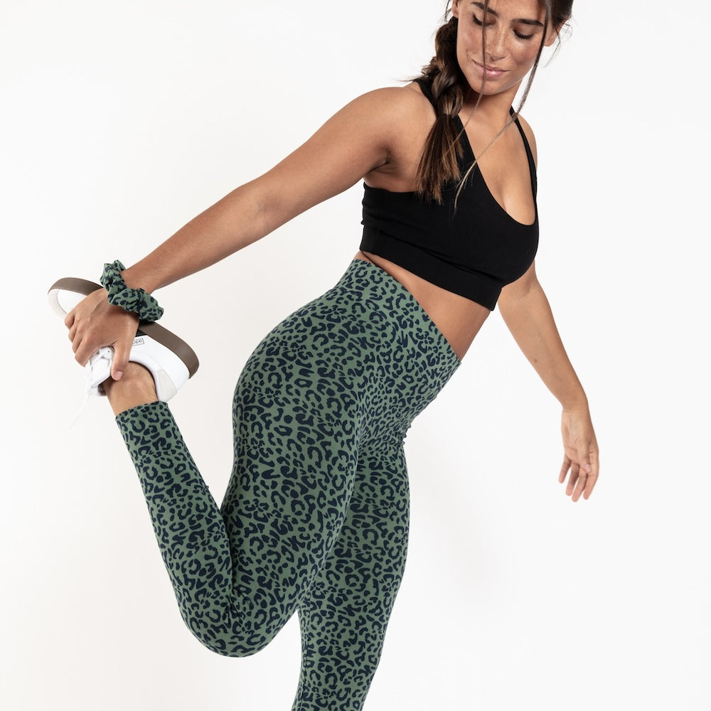 Woman wearing green leopard print leggings and black sports bra, stretching on a white background