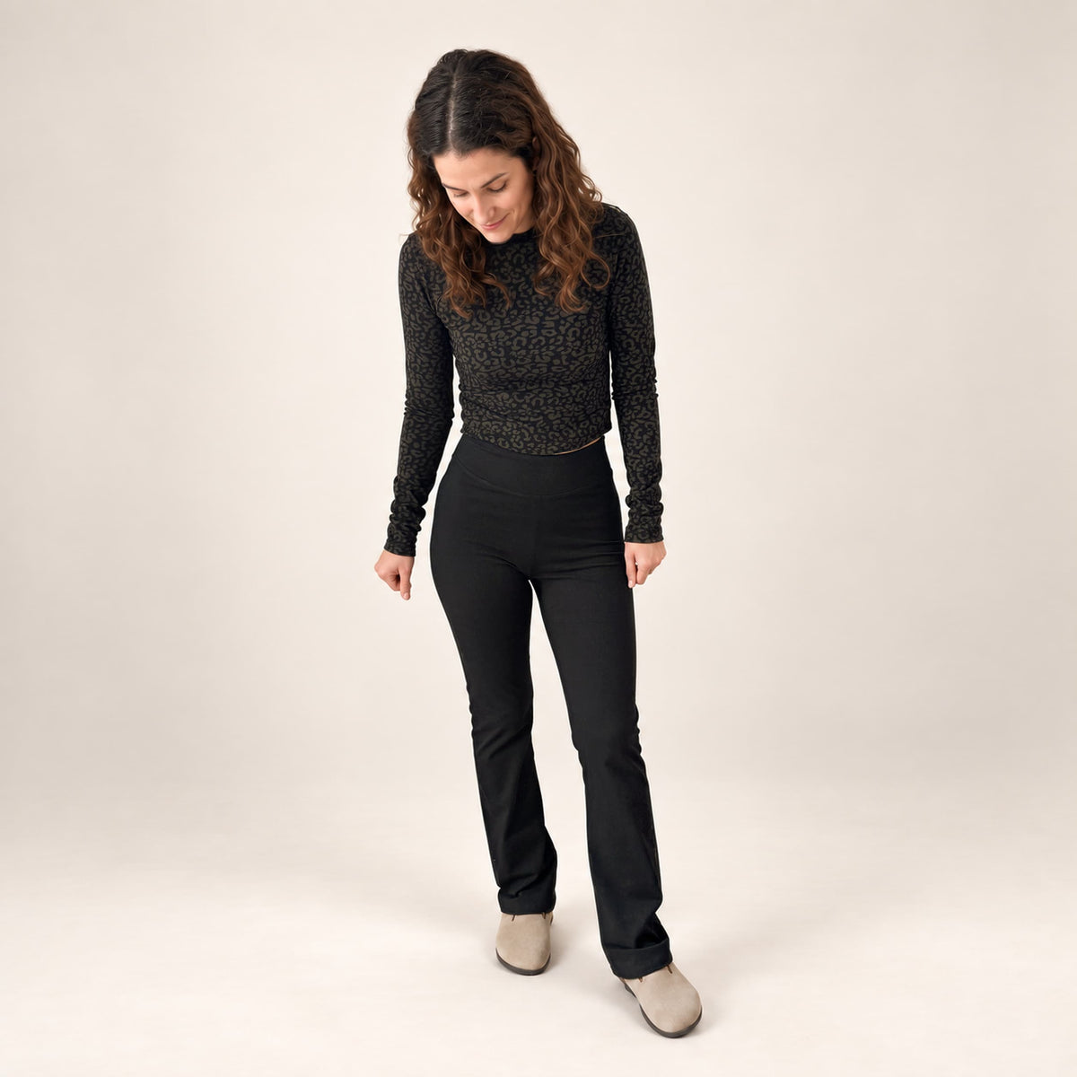 A woman modeling a black textured long-sleeve mock-neck top and high-waisted organic leggings flared leggings,, standing against a plain light beige studio background.